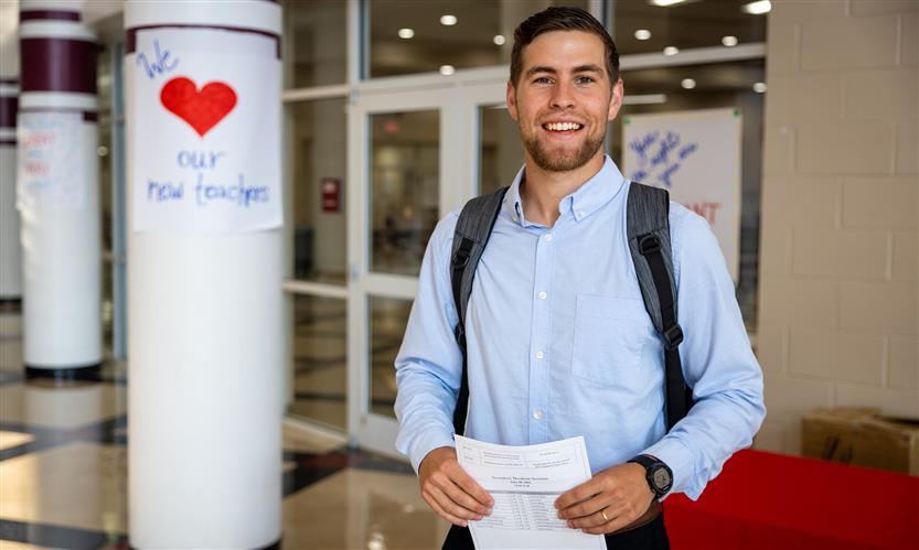 A man holding a piece of paper smiles while standing on front of a sign that reads \